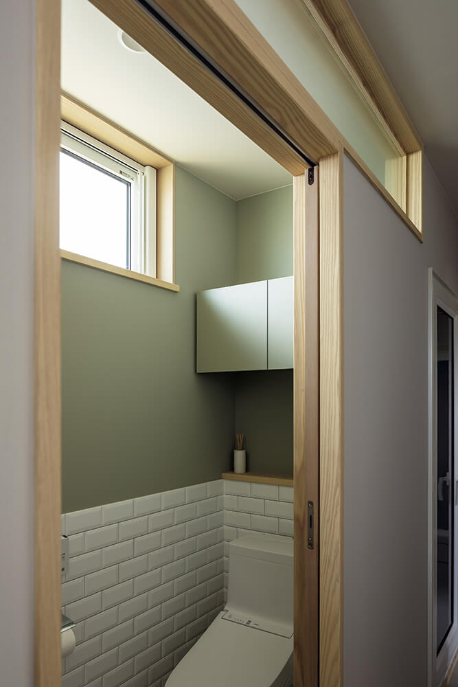 採光窓を間仕切り壁にも設け、廊下に光が抜けるよう仕上げたトイレ。淡いグレーのペイントと白いタイルの組み合わせも爽やか