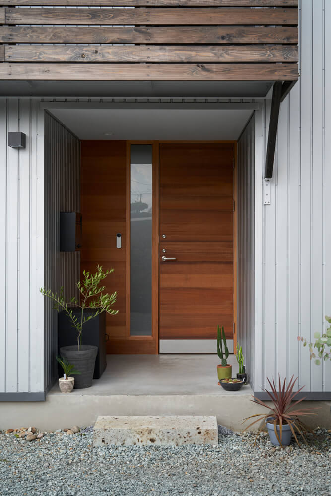 雨や雪対策として奥まらせた玄関。ガルバリウム鋼板のソリッドな印象の外壁に造作した木製玄関ドアがやわらかな印象を与える