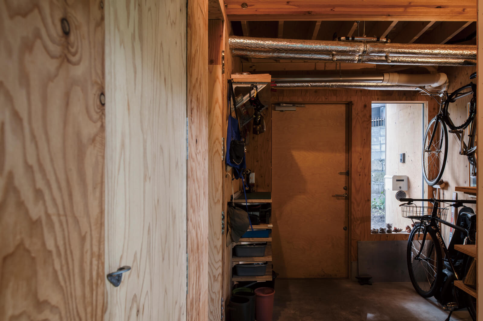 玄関まわりはKさん愛用の登山ギアが自作の棚にずらりと並ぶ。壁と合わせたラーチ合板でKさんがつくった建具のハンドルはクライミング用のハンガーを活用。露出配管も住まいの雰囲気になじんでいる