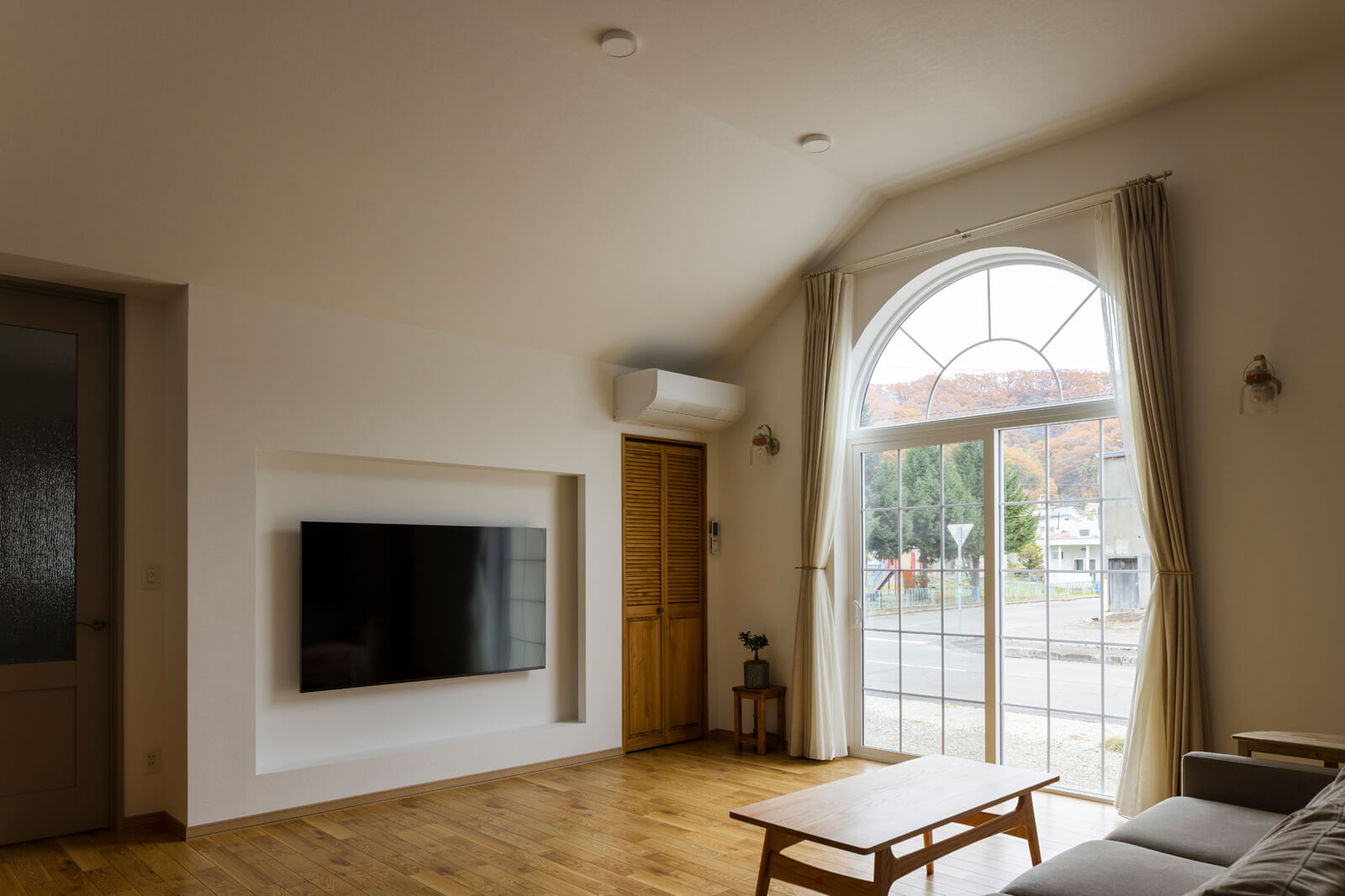 半円窓と掃き出し窓を組み合わせた大開口が開放感を演出するリビング。壁付けテレビの配線は壁の内側に収め、隣のクローゼット内にビデオデッキやオーディオ機器などを収納できる設計とした