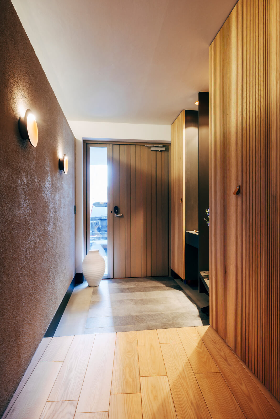 木質感にあふれた玄関。写真左側の壁は、ジョリパットの塗り壁仕様。凹凸のある壁面をウォールライトのやわらかい光が照らし、味わい深い雰囲気を醸す。リブ状に加工したナラ材の開き戸が目を引く造作の収納棚が廊下沿いに続く