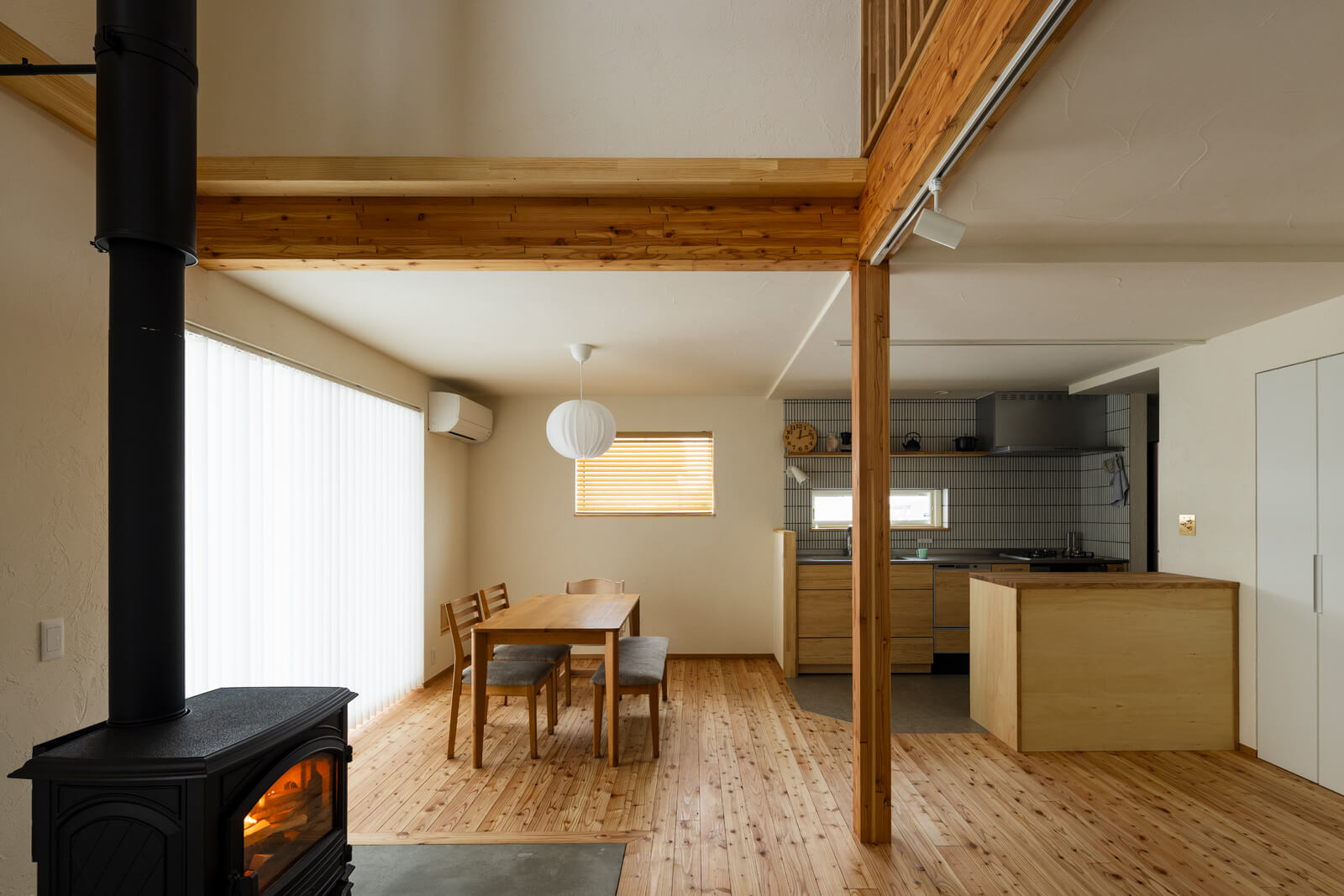 薪ストーブのある土間とダイニング、キッチンが一体となった空間。カラマツの床が空間全体に温かみを添える。リビングの大きな開口部にはバーチカルブラインドを採用し、光をやわらかく取り込みながら整った空間を演出している